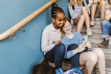Happy female friends sharing smart phone while sitting on staircase in school