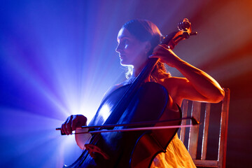 Cellist immersed in music, with striking halo of stage lights accentuating her silhouette on stage. Classic music in modern life. Concept of hobby and work, music festivals, concerts, symphony show.