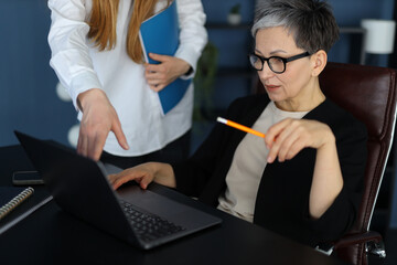 Two businesswomen in a modern office collaborate on a laptop, emphasizing successful teamwork and...