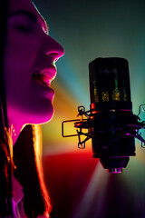 Profile portrait of female vocalist with studio microphone singing in stage lights casting colorful...