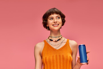 joyful young woman with short brunette hair holding soda can while posing on pink, summer beverage