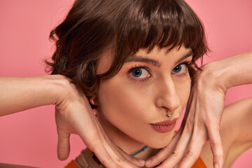 flirty young woman with nose piercing and short brunette hair pouting lips on pink background