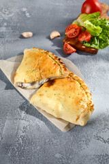 Calzone pizza with golden crust, served on a grey textured background, accompanied by fresh tomatoes and garlic