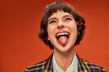 Playful female student in college uniform sticking out tongue, lively on orange background - 776111829