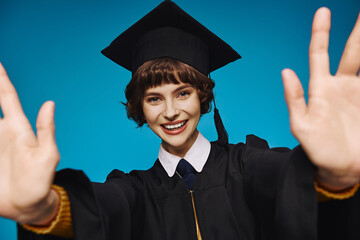 happy graduate college girl in academic cap with outstretched hands on blue backdrop, accomplishment