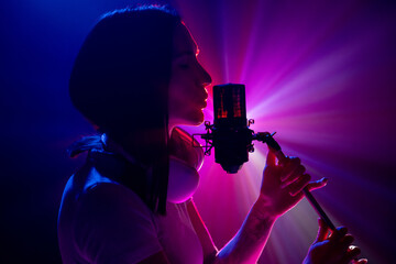 Side view portrait of silhouette of artist, young brunette woman singing at microphone with radiant colorful light show behind. Concept of art, work and hobby, music festivals, self expression. Ad