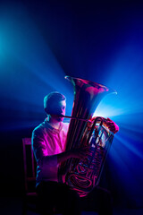 Portrait of talented musician seated, playing trumpet with soft blue-pink stage lights highlighted...