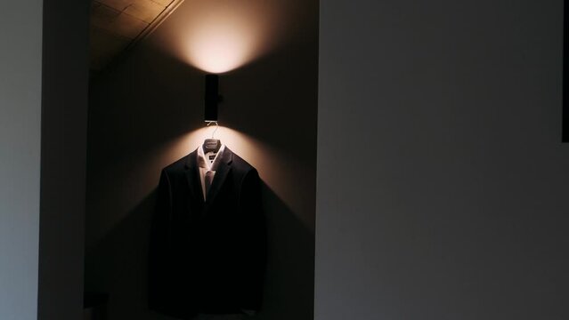 A man's jacket hangs on a hanger in the rays of light from a lamp in a dark room