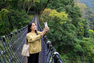 Woman walk along the suspension bridge and use mobile phone to take photo in the forest