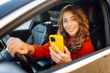 Portrait of cheerful woman looking phone behind steering wheel. The driver of the car uses a...