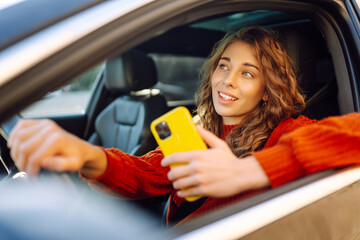 Portrait of cheerful woman looking phone behind steering wheel. The driver of the car uses a...