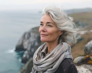 A woman with gray hair is standing on a rocky cliff overlooking the ocean. She is wearing a scarf and has a smile on her face