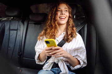 Young woman uses a smartphone while sitting in the back seat of a car. Female happy in car traveling on the road to destination. Technology, blogging, business concept. - obrazy, fototapety, plakaty