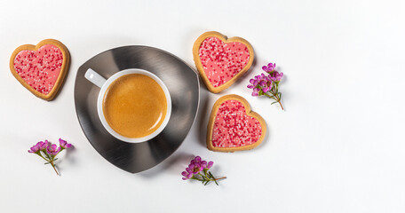 A cup of coffee on a saucer with three heart-shaped cookies in pink glaze and flower sprigs. Top...