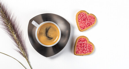A cup of coffee on a saucer with two heart-shaped cookies in pink glaze and a sprig of grass. Top view on a white background.
