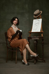 Elegant man in silky, old-fashion dress sitting by easel with self-portrait and drinking coffee...