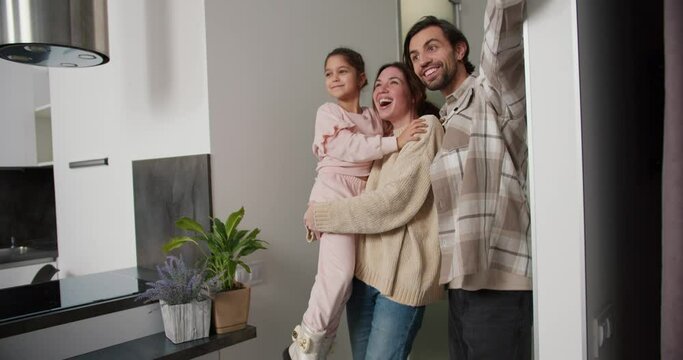 A happy brunette girl in a beige sweater carries her daughter in pink clothes in her arms and together with her boyfriend enters a modern apartment after moving. A happy family enters a new apartment