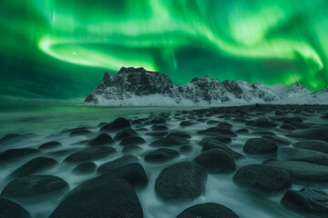 Low angle view of waves flowing over the rocks in the foreground with snow-capped mountains in the background at night. Aurora Borealis dancing over the beautiful rocky coastline of Uttakleiv beach