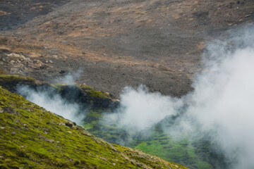 A cascading waterfall plunges down moss-covered mountains in a geothermal landscape