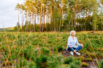 Naklejka na ściany i meble Elderly positive woman with grey hair enjoy beautiful sunny summer evening drinking sitting on meadow with woods on background.