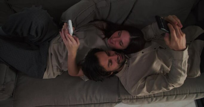A brunette guy with stubble and a girl in a beige T-shirt lie head to head on the sofa and watch something on their smartphones during their evening relaxation on the sofa