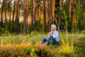 Beautiful senior woman sit at forest meadow at sunset and texting, admiring nature with pine woods on background.