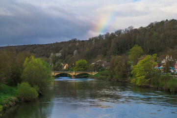 Obraz premium Regenbogen über Rundbogenbrücke an Fluss mit Uferböschung und Hochwasserschutzmauer in Neckartenzlingen.