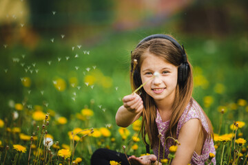 A cute little girl wearing headphones blowing dandelion fluff. - 776202427