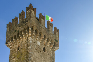Crenellated tower of the Scaliger Castle (13-14th centuries) with an Italian flag on top against clear blue sky, Sirmione, Brescia, Lombardy, Italy
