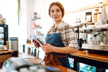 A young woman in an apron with a digital tablet at the bar counter of a coffee shop takes an order....