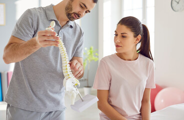 Portrait of male doctor physiotherapist or orthopedist showing to his woman patient model of spine in rehab clinic giving consultation about scoliosis or spinal problems during medical exam.