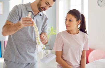 Portrait of male doctor physiotherapist or orthopedist showing to his woman patient model of spine...