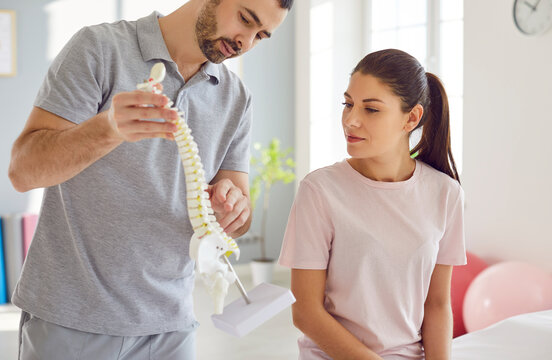 Portrait of male doctor physiotherapist or orthopedist showing to his woman patient model of spine in rehab clinic giving consultation about scoliosis or spinal problems during medical exam. - Powered by Adobe