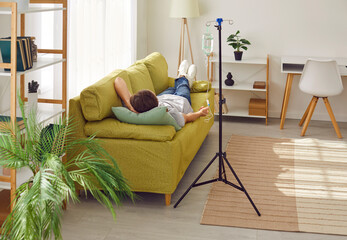 Back view of young man lying on the couch at home or in clinic while receiving IV drip infusion and...