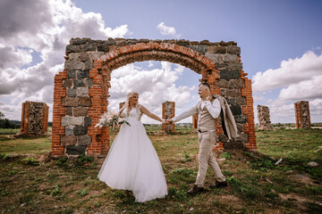 A newlywed couple holds hands under an archway amidst the ruins, surrounded by a landscape of sky...