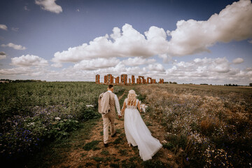 A couple walks hand in hand towards ancient ruins in a wildflower meadow under a dramatic sky,...