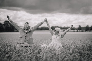 A jubilant bride and groom hold hands and raise their arms in triumph amidst a field of tall grass,...
