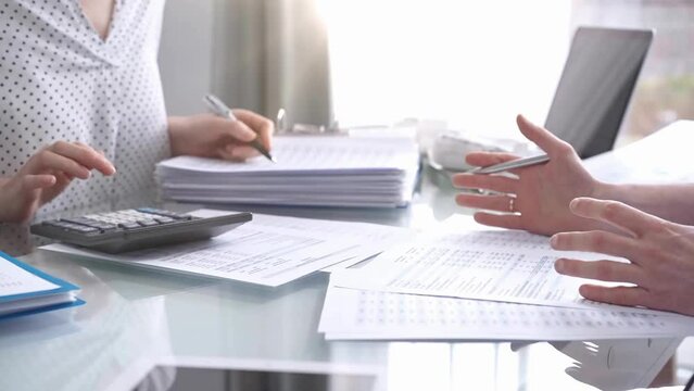 Business colleagues, accountants, or auditors are reviewing financial data in fair office. Two professionals are analyzing paper reports and using calculators, focusing on aspects of audit and taxes