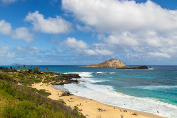 a beautiful spring landscape along the coast of Oahu with a sandy beach, blue ocean water, people relaxing, lush green trees and plants, rocks, blue sky and clouds in Honolulu Hawaii USA
