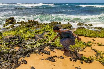 an African American woman holding a smooth white stone at the beach with silky brown sand and rocks covered in lush green algae at Sandy Beach in Honolulu Hawaii