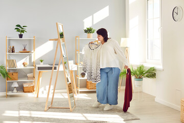 Overweight woman looking at mirror trying to choose clothes at home wardrobe. Plus size young woman holding hangers with blouses, thinking what to wear. Body positive concept - Powered by Adobe