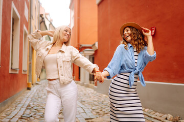 Jocund young ladies holding hands while walking down the street. Female tourists enjoys sights of the city on sunny day. Concept of lifestyle, fashion, travel.