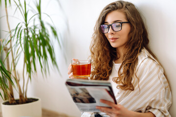 Young woman at home drinking coffee or tea and reading book or magazine. Lifestyle, relaxation and...