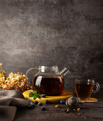 A glass teapot with black fragrant tea on a stand with a glass cup next to scattered blueberries...