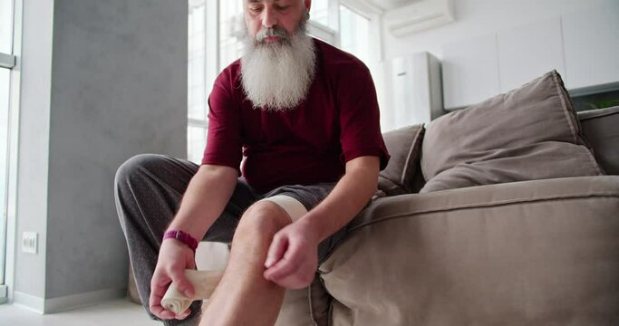 An elderly man with intact hair and a lush beard in a red T-shirt bandages his leg and knee with a bandage while sitting on a brown sofa in a modern apartment