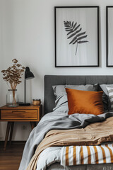 A modern bedroom with soft, grey bed linen and orange pillows on the gray fabric headboard against white walls. The room also features an industrial wooden bedside table holding some plants in vases.