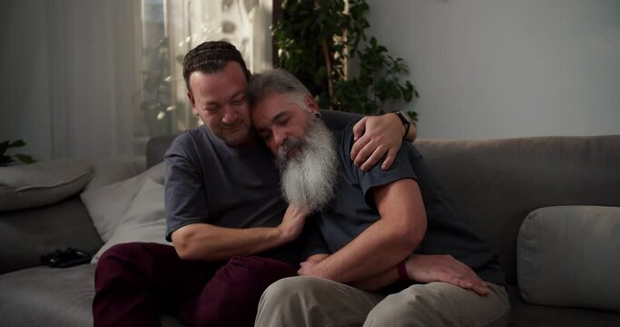 A middle-aged brunette man with stubble in a gray T-shirt hugs his sad boyfriend a man with gray hair color and a lush gray beard while sitting on a sofa in a modern apartment in the evening. Showing