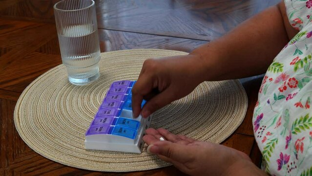 Hispanic woman placing pills into weekly pill box at kitchen table with glass of water