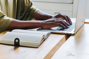 Close-up of a person's hands typing on a laptop keyboard, with a notebook beside on a wooden table,...