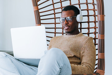 Relaxed African American Man Using Laptop with Headphones in Wicker Chair. A content man wearing...